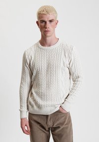 Maglione bianco lavorato a maglia con motivo a trecce, collo rotondo e polsini a costine. Indossato con pantaloni di velluto a coste color sabbia.