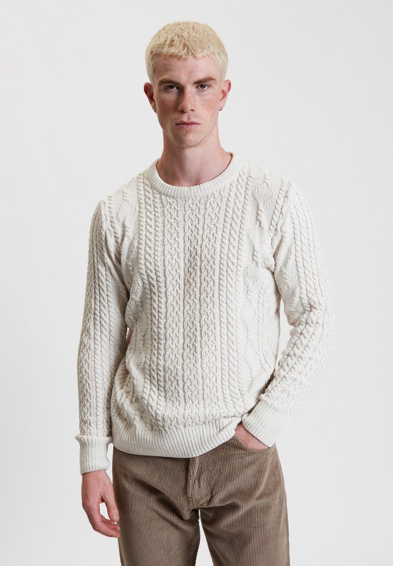Maglione bianco lavorato a maglia con motivo a trecce, collo rotondo e polsini a costine. Indossato con pantaloni di velluto a coste color sabbia.