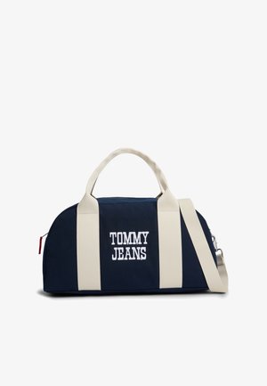 Navy blue duffel bag with white vertical straps, embroidered "TOMMY JEANS" text, top handles, and detachable shoulder strap.
