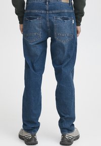 Denimu kopas, vidēji zilā krāsā, taisnas kājas griezums. Iespējas ietver divas aizmugurējās kabatas ar trīsstūra šuvēm un zīmola etiķeti uz jostas. Nēsāti ar pelēkiem apaviem.