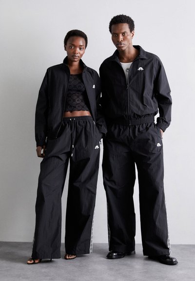 Vestes et pantalons de sport noirs, avec un design texturé, des tailles élastiques et des accents de logo blancs sur les manches et les côtés du pantalon.
