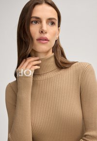 Beige Ripp-Turtleneck-Pullover mit taillierter Passform und weißen Logo-Akzenten an den Bündchen, verfügt über eine weiche Textur und lange Ärmel.