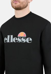 Felpa nera con colletto a girocollo a coste, grande logo "ellesse" in grigio e una grafica solare colorata in arancione e rosa sopra il logo.