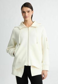 Witte zip-up hoodie met een grote capuchon, voorzakken en een zijzak met rits. Zachte textuur en een relaxte fit met een gesplitste zoom.