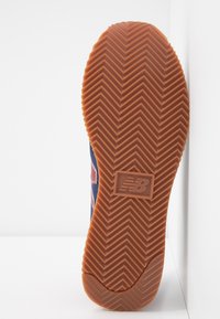 Semelle de basket en caoutchouc avec un motif en chevrons prononcé. La couleur est marron avec des accents rouges et bleu marine sur le dessus. Logo New Balance visible.