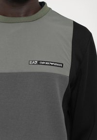 Cămașă cu mânecă lungă gri și neagră, cu un guler verde. Prezintă un design cu blocuri de culoare orizontale și un logo EA7 Emporio Armani.