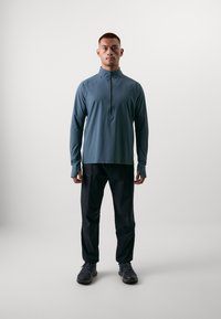 Lichte, lange mouw, rits-top in leisteenkleur, gecombineerd met zwarte, strakke broek. Beide zijn gemaakt van ademende stof.