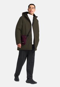 Olijfgroene waterdichte parka met een capuchon, ritsluiting en zijzakken, gedragen met zwarte broek en schoenen, vergezeld van een kleine bordeauxrode schoudertas.