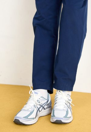 Weiße und hellblaue Sportschuhe mit grauen Akzenten und Schnürsenkeln, getragen mit blauen Hosen, vor einem gelben Hintergrund.