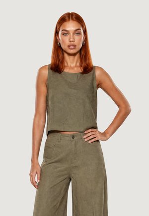 Donna con capelli rossi lisci che indossa un crop top verde oliva senza maniche e pantaloni larghi a vita alta, in piedi con una mano sul fianco.