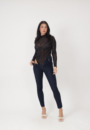 Kobieta poprawia rozpięte ciemne wąskie dżinsy, nosi przezroczystą czarną bluzkę z długim rękawem z wzorami oraz białe sandały na wysokim obcasie, stoi na białym tle.