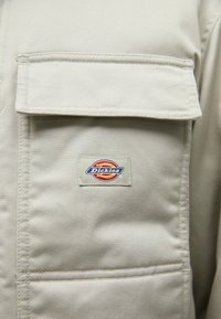 Μπεζ εργασιακή ζακέτα κατασκευασμένη από ανθεκτικό ύφασμα, με μία τσέπη στο στήθος και ένα ορθογώνιο λογότυπο "Dickies" με κόκκινες και μπλε λεπτομέρε�ιες.