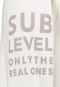 Witte sweatshirt met grote lichtgrijze tekst die leest "SUB LEVEL ONLY THE REAL ONES." Zacht, gestructureerd materiaal met een ontspannen pasvorm.
