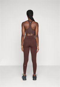 Mujer con trenzas, vistiendo un sujetador deportivo marrón oscuro y leggins, de pie de espaldas sobre un fondo claro y liso.