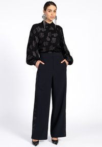 Black blouse with metallic pattern, long sleeves, and a collar paired with wide-leg navy trousers featuring satin side stripes. Black heeled shoes.