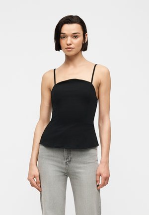 Jeune femme aux cheveux courts et foncés, portant un haut noir ajusté à fines bretelles et un jean gris clair sur un fond uni.