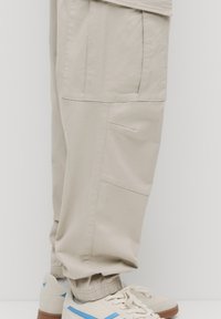 Beige cargo bukser med flere lommer, elastiske cuffs og en blød stoftekstur, parret med off-white sneakers med blå accenter.