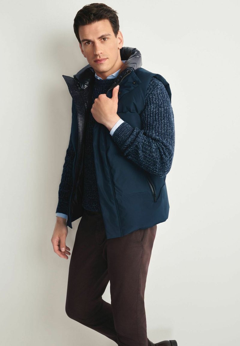 Granatowa kamizelka puchowa z wysokim kołnierzem, o matowym wykończeniu i kieszeniach na zamek, noszona na fakturowanym ciemnoniebieskim swetrze i brązowych spodniach.