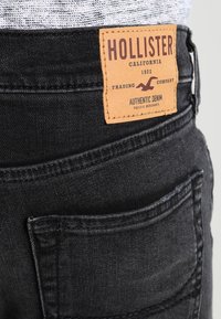 Jeans de mezclilla negra con una etiqueta de cuero que dice "HOLLISTER", con detalles de costura visibles y bolsillos traseros.