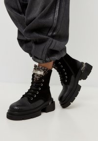Schwarze Knöchelstiefel aus strukturiertem Leder, mit runder Zehenform, dicker Sohle und metallischem Akzent. Getragen mit grauen umgeschlagenen Hosen, die ein Leopardenmuster-Futter zeigen.