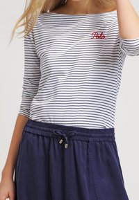 Långärmad randig topp i vitt med marinblå band, med en röd broderad logotyp. Matcher med en marinblå dragsko-kjol i slät textur.