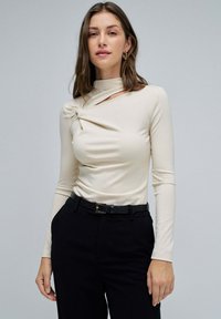 Top beige de manga larga con un detalle retorcido en el escote, combinado con pantalones negros y un cinturón negro texturizado. Tela suave y elástica.