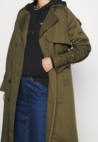 Olivgrön trenchcoat med dubbelknäppt design, stor krage och knapprygg, bärs över en svart hoodie och blå jeans.