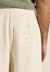 Beige bomuldsbukser med en textureret overflade, der har en baglomme og et mærke med teksten "LA MAISON AMV" i grønt på en hvid baggrund.
