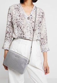Mujer vestida con una blusa de estampado de piel de serpiente y pantalones blancos, llevando un bolso cruzado de Michael Kors en color plateado con correa de cadena.