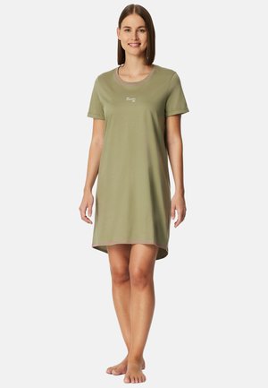 Jonge vrouw staat blootsvoets, draagt een olijfgroene jurk met korte mouwen, roze afwerking en kleine witte tekst op de borst.