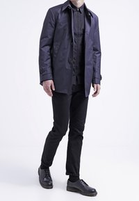 Imperméable marine au design élégant, avec des boutons et un col ; porté par-dessus une chemise noire et un pantalon noir, assorti à des chaussures noires.