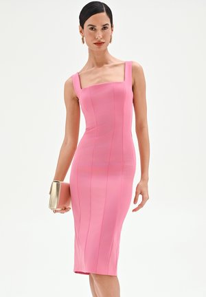 Femme portant une robe midi rose ajustée avec larges bretelles, tenant une pochette dorée et portant des boucles d'oreilles créoles dorées sur un fond uni.