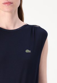 Granatowa, bezrękawnikowa bluzka z delikatnym marszczeniem na ramieniu oraz zielono-białym logo Lacoste wyszytym po lewej stronie klatki piersiowej.