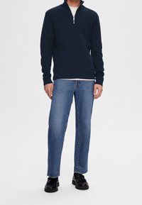 Laivastonsininen neulepusero puolivetoketjukauluksella, yhdistetty vaaleansinisiin suoralahkeisiin farkkuihin ja mustiin kenkiin. Yksinkertainen, klassinen muotoilu.