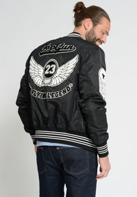 Zwarte bomberjack met geborduurde vleugels en het nummer "23" op de achterkant. Voorzien van gestreepte manchetten en onderrand. Draagt over een spijkerbroek.