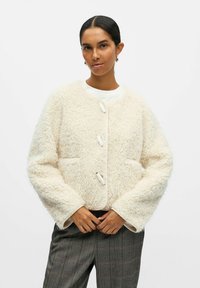 Giacca texturizzata color crema con scollo rotondo, tre bottoni e tasche laterali; indossata sopra una maglia bianca e pantaloni in plaid grigi.