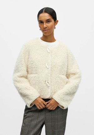 Veste texturée crème avec un col rond, trois boutons et des poches latérales ; portée sur un haut blanc et un pantalon à carreaux gris.