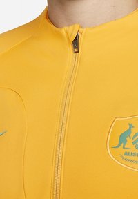 Gul atletisk zip-up jakke lavet af glat stof. Har et kænguru logo og ordet "AUSTRALIEN" i blå. Lynlås med sølvhardware.