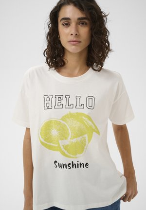 Kobieta o kręconych brązowych włosach, ubrana w oversize'owy biały T-shirt z nadrukiem żółtych plasterków cytryny i napisem „HELLO Sunshine” z przodu.