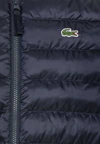 Chaqueta acolchada de un oscuro color navy con cierre de cremallera, diseño de acolchado horizontal y un pequeño logo de cocodrilo verde en el lado izquierdo del pecho.