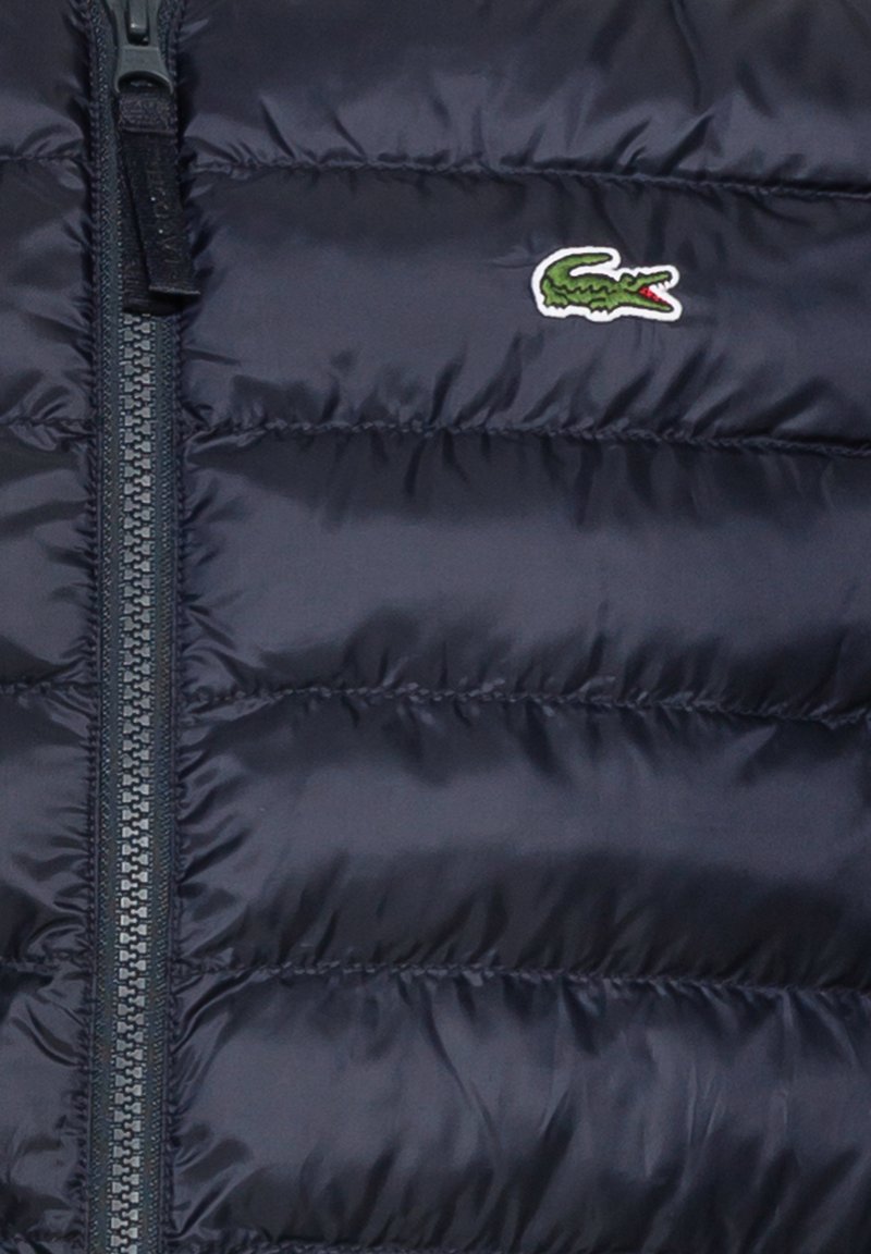Doudoune bleu marine foncé avec fermeture éclair, design matelassé horizontal et un petit logo crocodile vert sur le côté gauche de la poitrine.