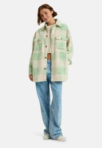 Geverifieerde oversized jas in crème en mintgroen, met twee voorkruiszakken, knopen en een textielachtige stof. Gecombineerd met lichte denim jeans.