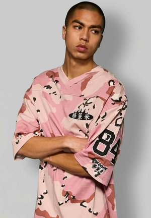 Junger Mann mit verschränkten Armen, der ein rosa-braunes Hemd mit Tarnmuster trägt, auf dem die Zahl "84" und asiatische Schriftzeichen auf Ärmel und Brust aufgedruckt sind.