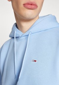 Felpa blu chiaro con cappuccio regolabile e un piccolo logo ricamato con dettagli rossi e blu scuro nell'area del petto sinistro. Tessuto morbido.