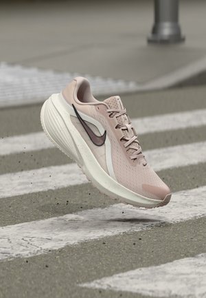 Jasnoróżowy but do biegania marki Nike unoszący się w powietrzu nad przejściem dla pieszych, widok z boku z czarnym logo Swoosh i teksturowaną podeszwą.