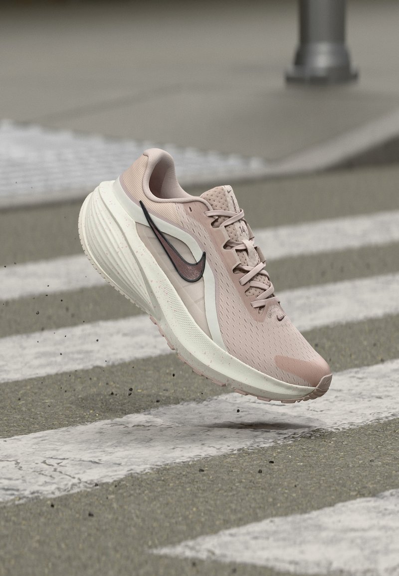 Ροζ Nike αθλητικό παπούτσι στον αέρα πάνω από διάβαση πεζών, που δείχνει πλευρική όψη με μαύρο λογότυπο Swoosh και ανάγλυφη σόλα.