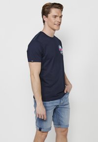 Tumši zila kokvilnas t-kreklu ar īsām piedurknēm un apaļu kakla izgriezumu. Priekšā ir krāsains grafiskais dizains un kontrastējošs šuvums.