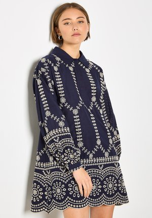 Joven con cabello corto y castaño que lleva un vestido azul marino con bordados florales blancos, de pie contra un fondo claro y liso.