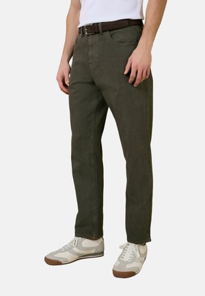 Egy férfi sötétzöld egyenes szárú nadrágot visel barna övvel, fehér tornacipővel és fehér ujjatlan felsővel, egyszínű háttér előtt áll.