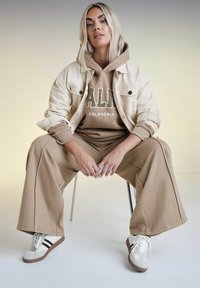 Beige oversized jacka över en brun hoodie med texten "CALIFORNIA", i kombination med matchande lösa byxor och vita sneakers med svarta ränder.